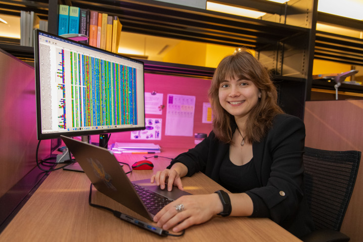 A woman with brown hair sits in front of two computer screens, filled with colorful data. 